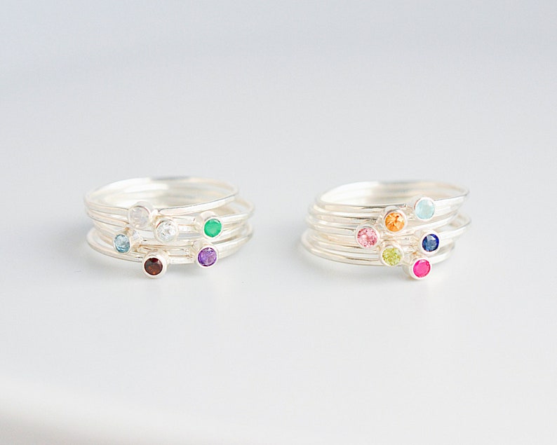 May include: Set of five silver stacking rings with colorful gemstones. The rings feature a variety of colors, including red, green, blue, purple, and white.