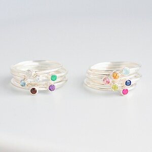 May include: Two stacks of silver rings, each with multiple bands. Each ring features a small, colorful gemstone. The gemstones include blue, clear, green, purple, and red. The other stack has pink, orange, light blue, dark blue, yellow, and magenta gemstones.