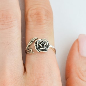 May include: A silver-toned ring featuring a detailed rose design. The rose is the focal point, with delicate petals and leaves wrapping around the finger. The ring is worn on a finger, showcasing the floral jewelry.