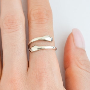 May include: A silver ring with two hands clasped together, forming a circle. The ring is worn on a finger.