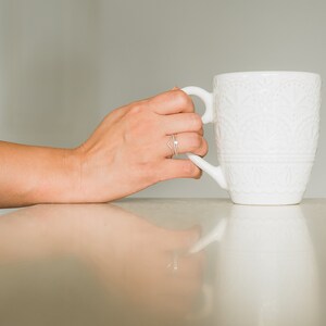 May include: A white ceramic mug with an embossed design is being held by a hand wearing a silver ring.
