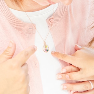 May include: A woman wearing a pink cardigan with a gold necklace featuring a small, oval-shaped pendant with colorful gemstones. The pendant is hanging on a delicate chain.
