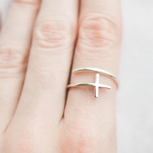 Könnte beinhalten: Ein silberner Ring mit einem zarten Kreuzdesign. Der Ring ist ein Doppelband, wobei ein Band um den Finger gewickelt ist und das andere Band das Kreuz trägt.