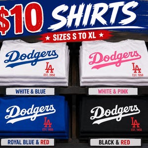May include: Four stacks of folded t-shirts featuring the Dodgers logo and LA emblem with EST. 1958 text. The color combinations include white with blue, white with pink, royal blue with red, and black with red. The display includes labels for each.