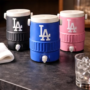 Puede incluir: Tres enfriadores de bebidas aislados en negro, azul y rosa, cada uno con una tapa blanca y el logotipo blanco "LA". Los enfriadores tienen un grifo para dispensar bebidas y están sobre una encimera oscura.