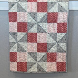 May include: A handmade quilt with a patchwork design. The quilt features squares and triangles in shades of red, pink, and gray. The squares have a floral pattern, and the triangles have a solid color. The quilt is hanging on a white bar.