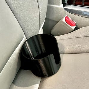 May include: A black, cylindrical cup holder insert with a smooth finish and a vertical cutout along one side, designed to fit securely between the seat cushion and the center console of a vehicle interior with light-colored leather upholstery.