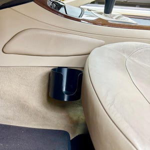 May include: A black, cylindrical car cup holder installed in a vehicle. The cup holder is mounted on the side of the center console, near the passenger seat. The interior of the car has tan leather seats and a beige carpet.