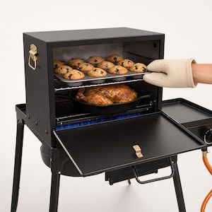 Puede incluir: Un horno portátil negro para exteriores con puerta abatible, que cuenta con una rejilla metálica con un molde para doce muffins horneados y una rejilla inferior con un pollo asado en una sartén de hierro fundido, con llamas azules visibles.