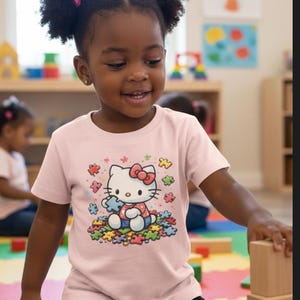 May include: A young child wearing a pale pink t-shirt with a Hello Kitty graphic. The graphic depicts Hello Kitty holding a puzzle piece, surrounded by colourful puzzle pieces. The child is smiling and playing with wooden blocks.