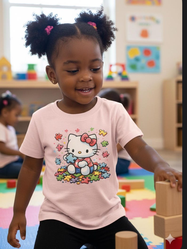 May include: A young child wearing a pale pink t-shirt with a Hello Kitty graphic. The graphic depicts Hello Kitty holding a puzzle piece, surrounded by colourful puzzle pieces. The child is smiling and playing with wooden blocks.