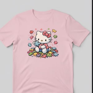 May include: A pale pink t-shirt with a cartoon illustration of Hello Kitty holding a puzzle piece. She is surrounded by colourful puzzle pieces. Hello Kitty wears a red bow, a red and white polka dot dress, and blue denim dungarees.
