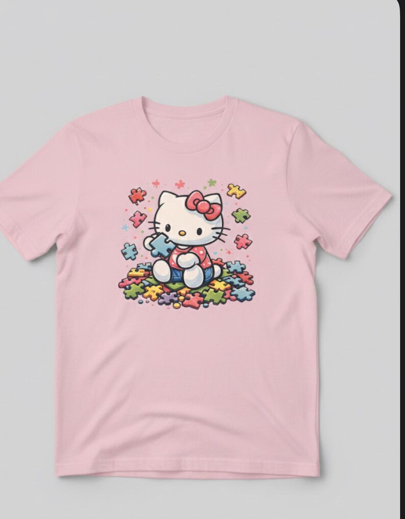 May include: A pale pink t-shirt with a cartoon illustration of Hello Kitty holding a puzzle piece. She is surrounded by colourful puzzle pieces. Hello Kitty wears a red bow, a red and white polka dot dress, and blue denim dungarees.