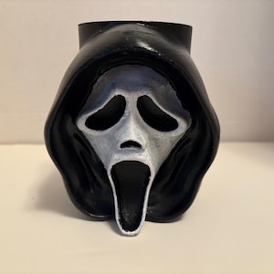 May include: A black and white Scream mask, a popular horror movie icon. The mask has a white face with black eye holes and a wide open mouth. The mask is set against a plain background.