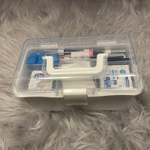 May include: A clear plastic toolbox with a white handle and latch, filled with various items. The contents include blue foam pieces, pens, and packets of wipes. The toolbox is sitting on a gray, fuzzy surface.
