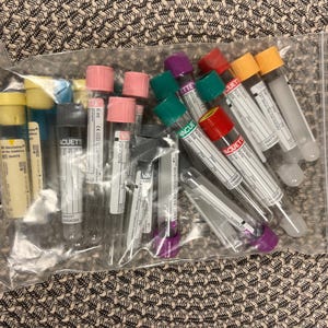 May include: A clear plastic bag filled with various medical test tubes. The tubes have colored caps in yellow, pink, green, red, and orange. Some tubes have labels with text. The bag is on a patterned surface.