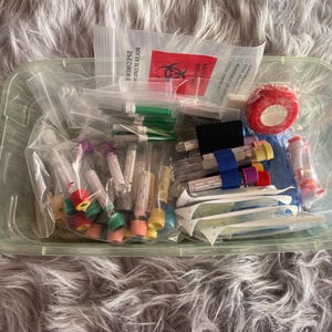 May include: A clear plastic container filled with medical supplies, including vials with colored caps, needles, bandages, and a red biohazard bag. The items are packaged in clear plastic bags.