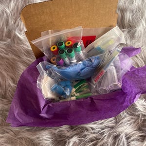 May include: A cardboard box filled with medical supplies, including vials with colorful caps, blue gloves, and other tools. The items are packaged in clear plastic bags and surrounded by purple tissue paper.