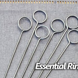 May include: Six silver metal skewers with a ring at the top. The skewers are arranged in a fan shape on a gray fabric background. The text "Essential Ring" is printed below the skewers.
