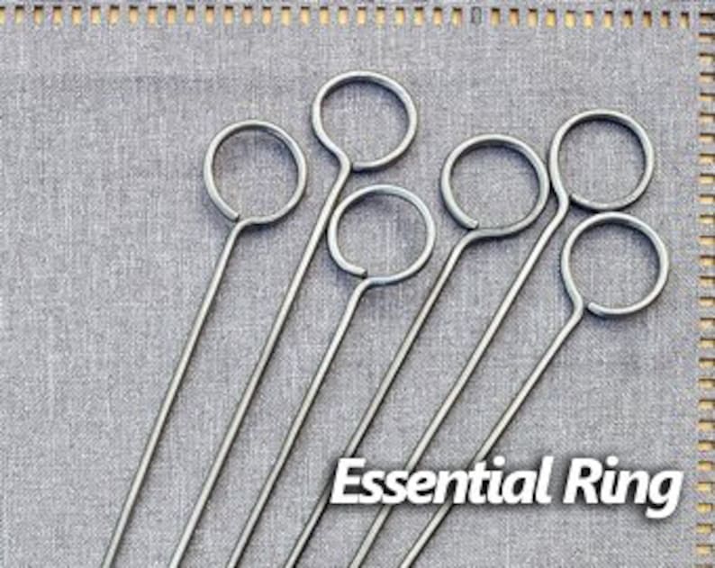 May include: Six silver metal skewers with a ring at the top. The skewers are arranged in a fan shape on a gray fabric background. The text "Essential Ring" is printed below the skewers.
