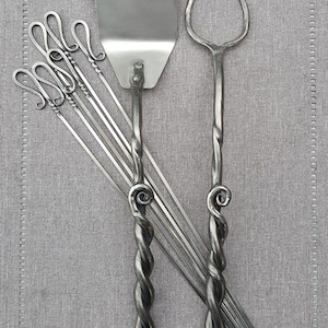 May include: A set of forged steel grilling tools, including a spatula, a fork, and six skewers. The tools have twisted handles and are made in a rustic style.