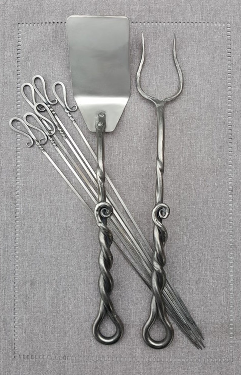 May include: A set of forged steel grilling tools, including a spatula, a fork, and six skewers. The tools have twisted handles and are made in a rustic style.