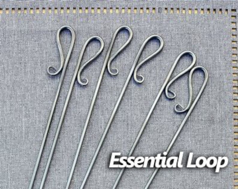 May include: Six silver metal skewers with a decorative loop at the top. The skewers are arranged in a row on a gray fabric background. The text "Essential Loop" is printed below the skewers.