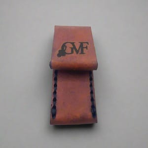 May include: A brown leather wallet with the letters "GMF" printed on the front. The wallet is folded open, revealing the interior. The edges are stitched with dark blue thread. The wallet is made of a textured leather material.
