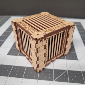 May include: A laser-cut wooden miniature cage with a brick-like exterior and vertical bars. The top features horizontal slats. The cage is a light brown color with darker brown accents, perfect for tabletop gaming or model building.