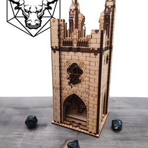 May include: A wooden dice tower in the shape of a castle with a bull logo. The tower is made of light brown wood and has a detailed design. Three dark blue dice are scattered around the tower.