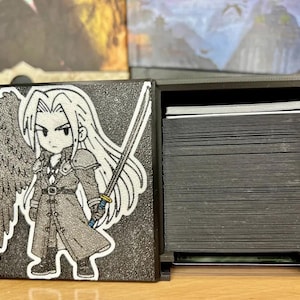 May include: A black card storage box with a detailed illustration of a character with long white hair, a sword, and a single wing. The box is open, revealing a stack of black cards. The box is made of a textured material.
