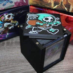 May include: Black card storage box with a removable lid featuring a green and white cartoon character with a skull face. The lid is decorated with a black background and the character is running with a bag over its shoulder.