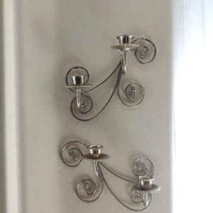 May include: Two silver wall-mounted candle holders with a decorative scroll design. Each holder has three candle cups.