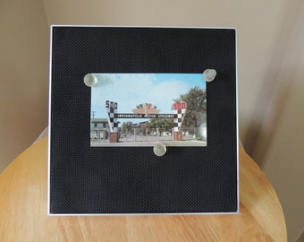May include: A black and white postcard with a photo of the Indianapolis Motor Speedway entrance. The entrance has a checkered flag design and the words "Indianapolis Motor Speedway" are printed on the archway. The postcard is displayed on a black mesh board with white push pins.
