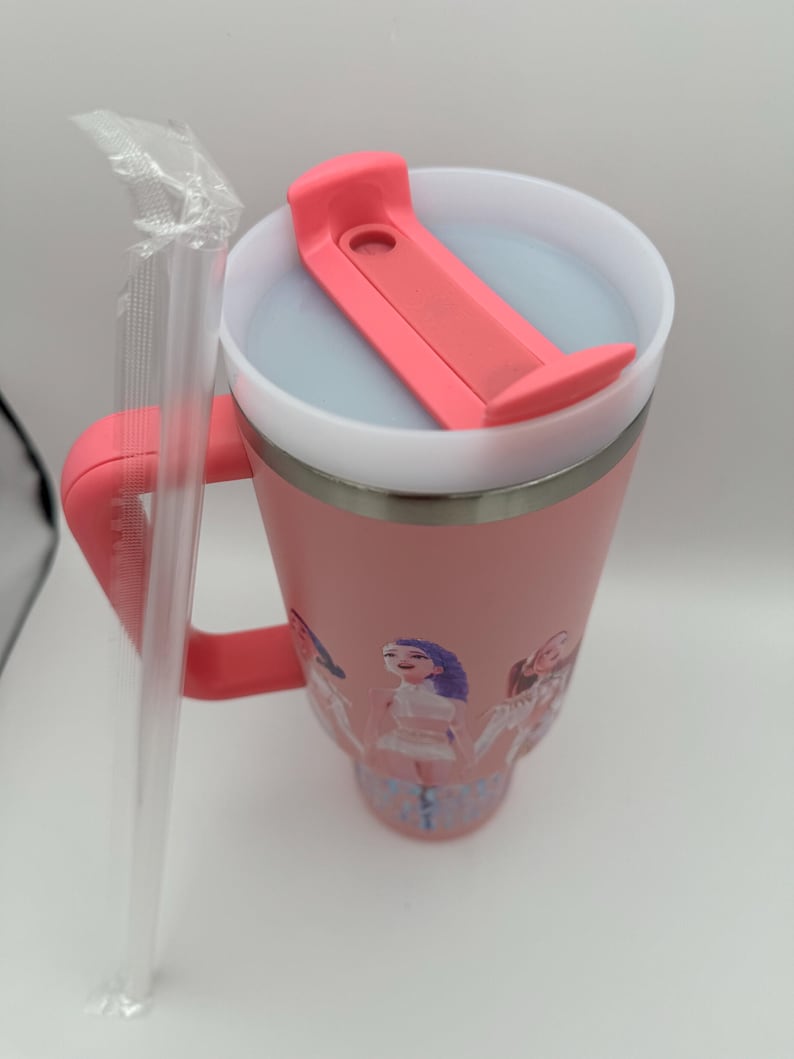 May include: A pink insulated tumbler with a handle and a lid featuring a pink flip-top closure. The tumbler has a silver rim and a design of cartoon figures. A clear plastic straw is included.
