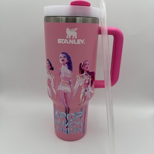 May include: Pink Stanley tumbler with a white lid and handle. The tumbler features a graphic of three cartoon figures and the text "KPOP DEMON HUNTERS". A clear plastic straw is included.