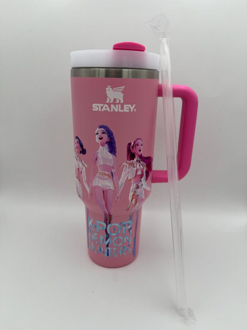 May include: Pink Stanley tumbler with a white lid and handle. The tumbler features a graphic of three cartoon figures and the text "KPOP DEMON HUNTERS". A clear plastic straw is included.