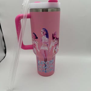 May include: Pink insulated tumbler with a white lid, pink handle, and a clear straw. The tumbler features a graphic of three cartoon women and the text "K-POP DEMON HUNTERS" in blue.