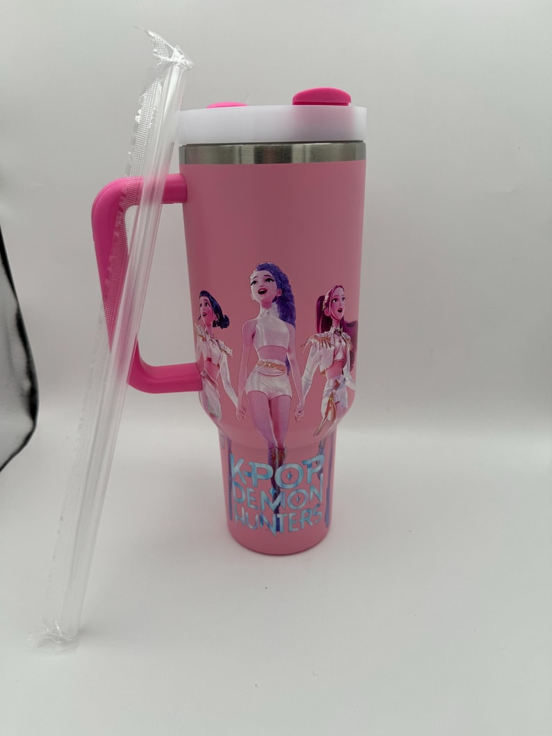 May include: Pink insulated tumbler with a white lid, pink handle, and a clear straw. The tumbler features a graphic of three cartoon women and the text "K-POP DEMON HUNTERS" in blue.