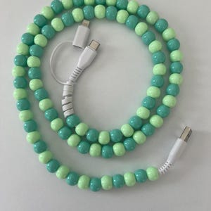 以下が含まれることがあります： ビーズデザインの装飾的なUSB充電ケーブル。ケーブルは、薄緑色とターコイズ色のビーズが交互に並んでいます。白いケーブルは、一方の端にUSB-Aコネクタ、もう一方の端にUSB-CおよびLightningコネクタを備えています。