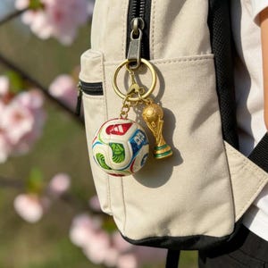 May include: A gold-colored keychain with two charms: a miniature soccer ball and a trophy. The soccer ball charm is white with colorful geometric designs. The trophy charm is gold with green and gold accents. The keychain is attached to a beige backpack.