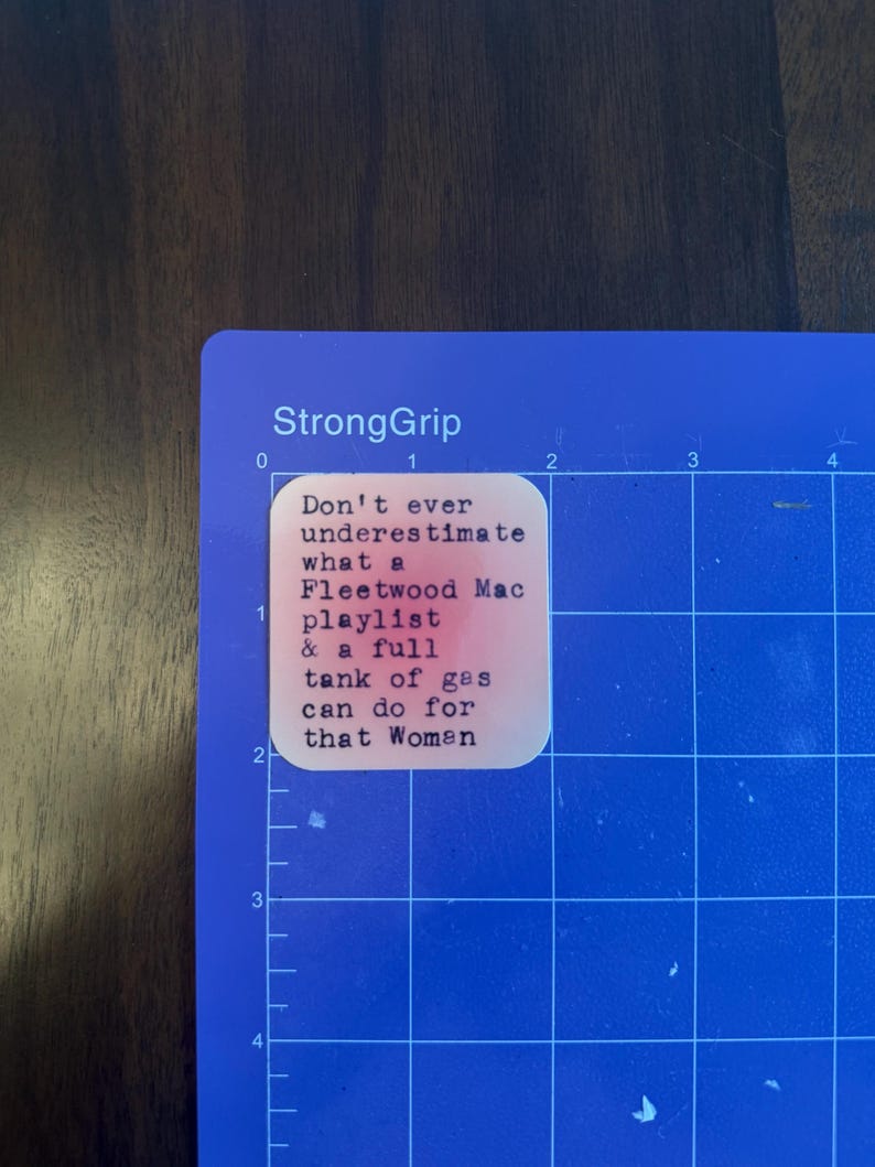 May include: A blue cutting mat with a white sticker that reads "Don't ever underestimate what a Fleetwood Mac playlist & a full tank of gas can do for that Women."