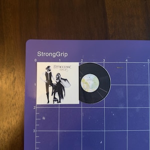 May include: A miniature vinyl record and album cover featuring the Fleetwood Mac album "Rumours." The album cover shows a black and white image of two people. The record is black with a silver label.