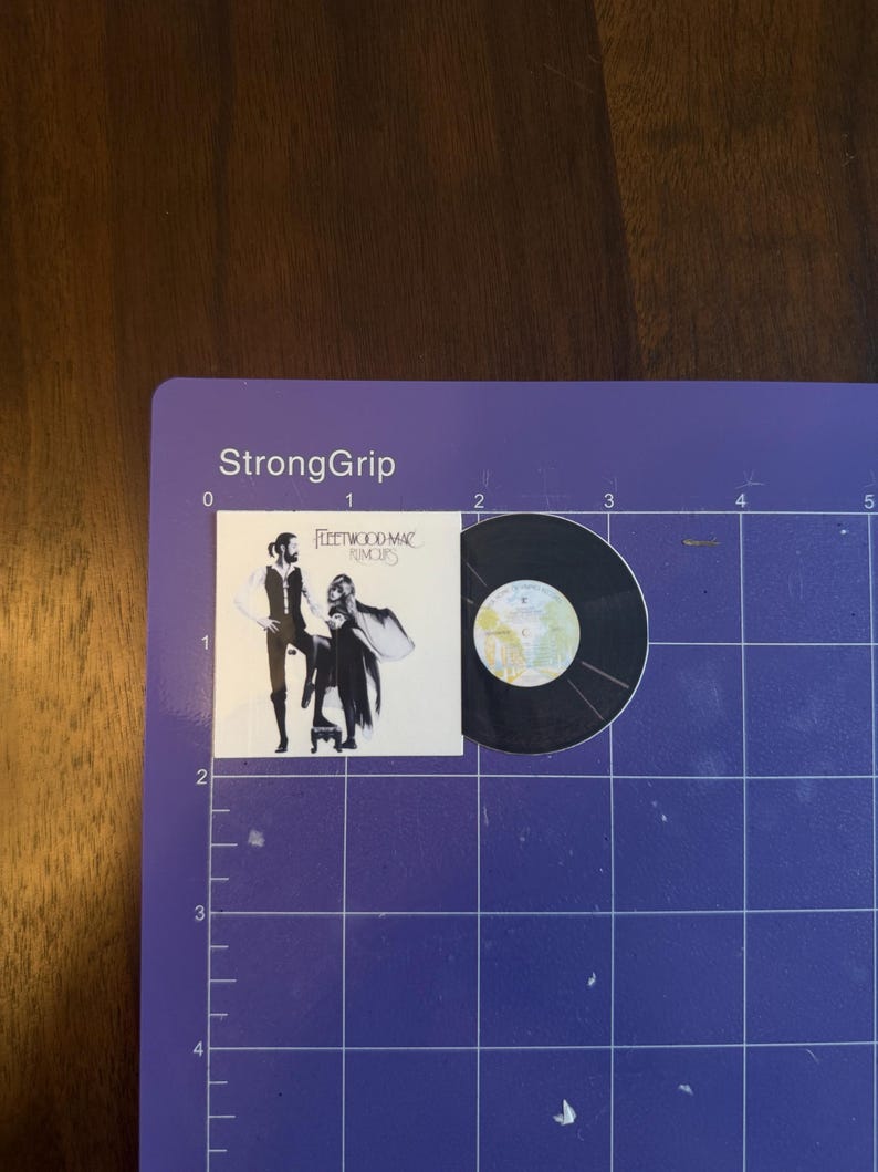 May include: A miniature vinyl record and album cover featuring the Fleetwood Mac album "Rumours." The album cover shows a black and white image of two people. The record is black with a silver label.