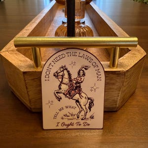 Puede incluir: Una bandeja de madera con asas doradas sostiene velas y una tarjeta decorativa. La tarjeta presenta a una vaquera montando a caballo con el texto "I DON'T NEED THE LAWS OF MAN TELL ME WHAT I OUGHT TO DO."