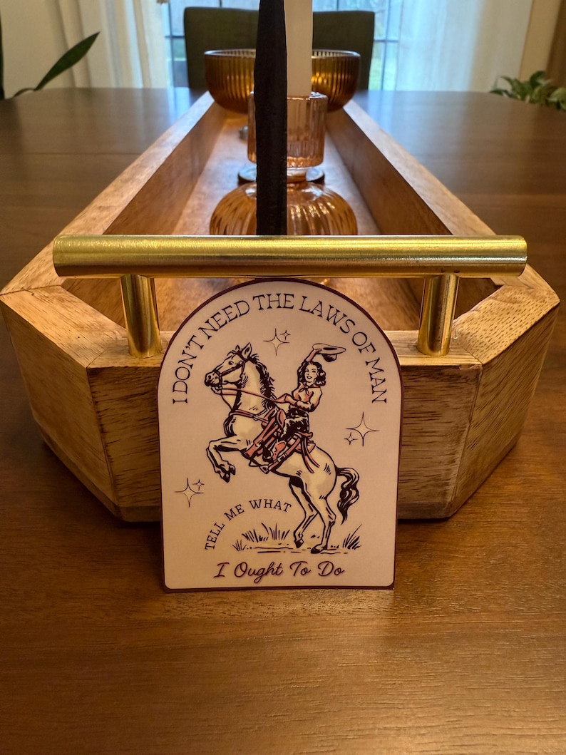 Puede incluir: Una bandeja de madera con asas doradas sostiene velas y una tarjeta decorativa. La tarjeta presenta a una vaquera montando a caballo con el texto "I DON'T NEED THE LAWS OF MAN TELL ME WHAT I OUGHT TO DO."