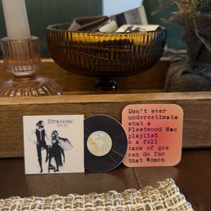 May include: A collection of Fleetwood Mac themed items. Includes a square album cover print, a vinyl record, and a coaster with text that reads "Don't ever underestimate what a Fleetwood Mac playlist & a full tank of gas can do for that Woman."