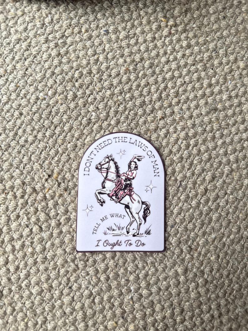 Puede incluir: Una pegatina rectangular con fondo blanco y borde granate. La pegatina presenta a una mujer montando un caballo encabritado, con el texto "I don't need the laws of man, tell me what I ought to do."