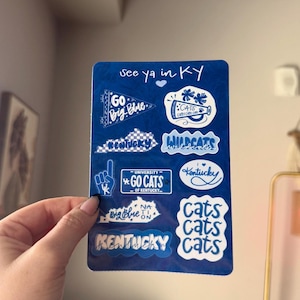 May include: A sheet of blue stickers with white text and graphics. The stickers feature phrases like "See ya in KY", "Go Big Blue", "Wildcats", and "Go Cats". The stickers are held by a hand.