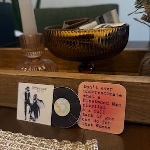 May include: A decorative wooden tray holds a brown glass bowl, a pink candle holder with a white candle, and two coasters. One coaster features a Fleetwood Mac album cover, and the other reads, "Don't ever underestimate what a Fleetwood Mac playlist & a full tank of gas can do for that Woman."